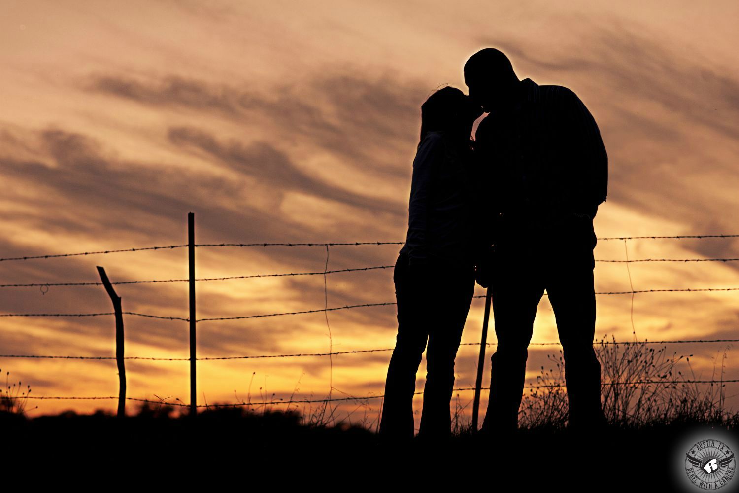silhouette of engagement couple kissing  with barbed wire fence and big orange sky after sunset in background.  It is a somewhat cloudy day and clouds are blue grey in the background